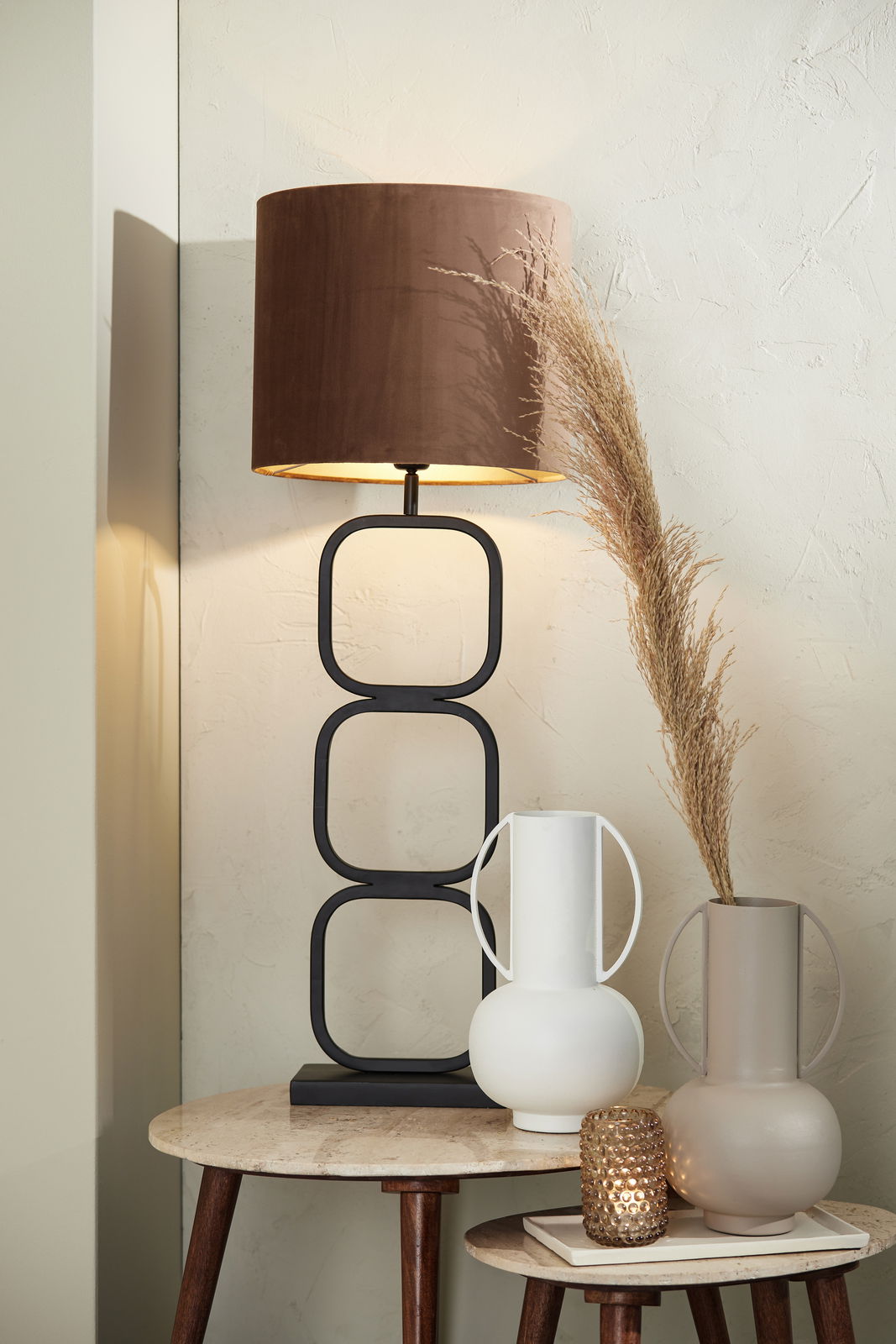 Moderne Tischlampe mit braunem Schirm und geometrischem Metallfuß, seitliche Perspektive, auf einem Tisch mit zwei dekorativen Vasen und einem Teelichthalter.