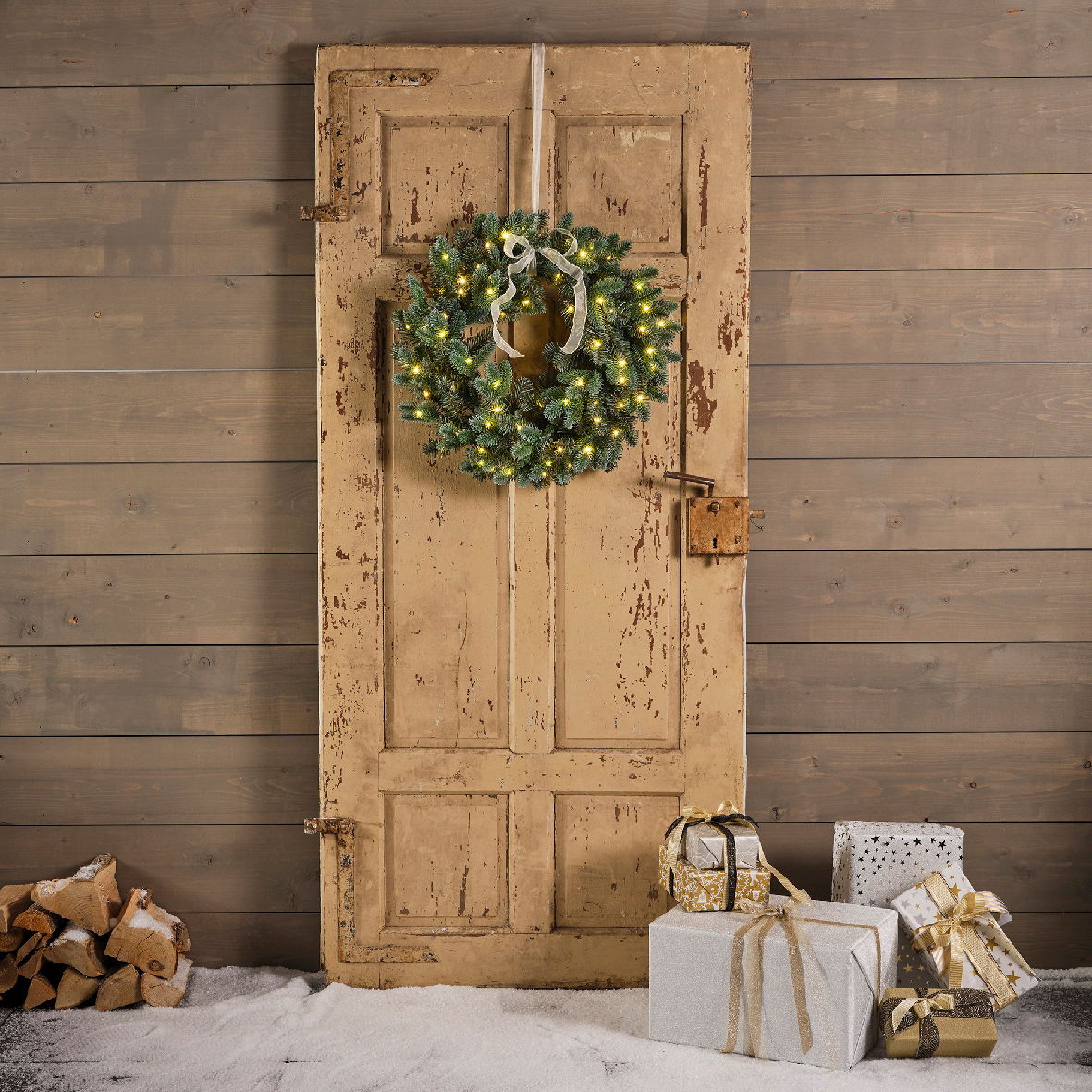 Weihnachtskranz mit Lichtern an einer alten Holztür, umgeben von Geschenken und Holzscheiten, aus frontaler Perspektive.
