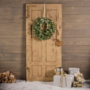 Weihnachtskranz mit Lichtern an einer alten Holztür, umgeben von Geschenken und Holzscheiten, aus frontaler Perspektive.