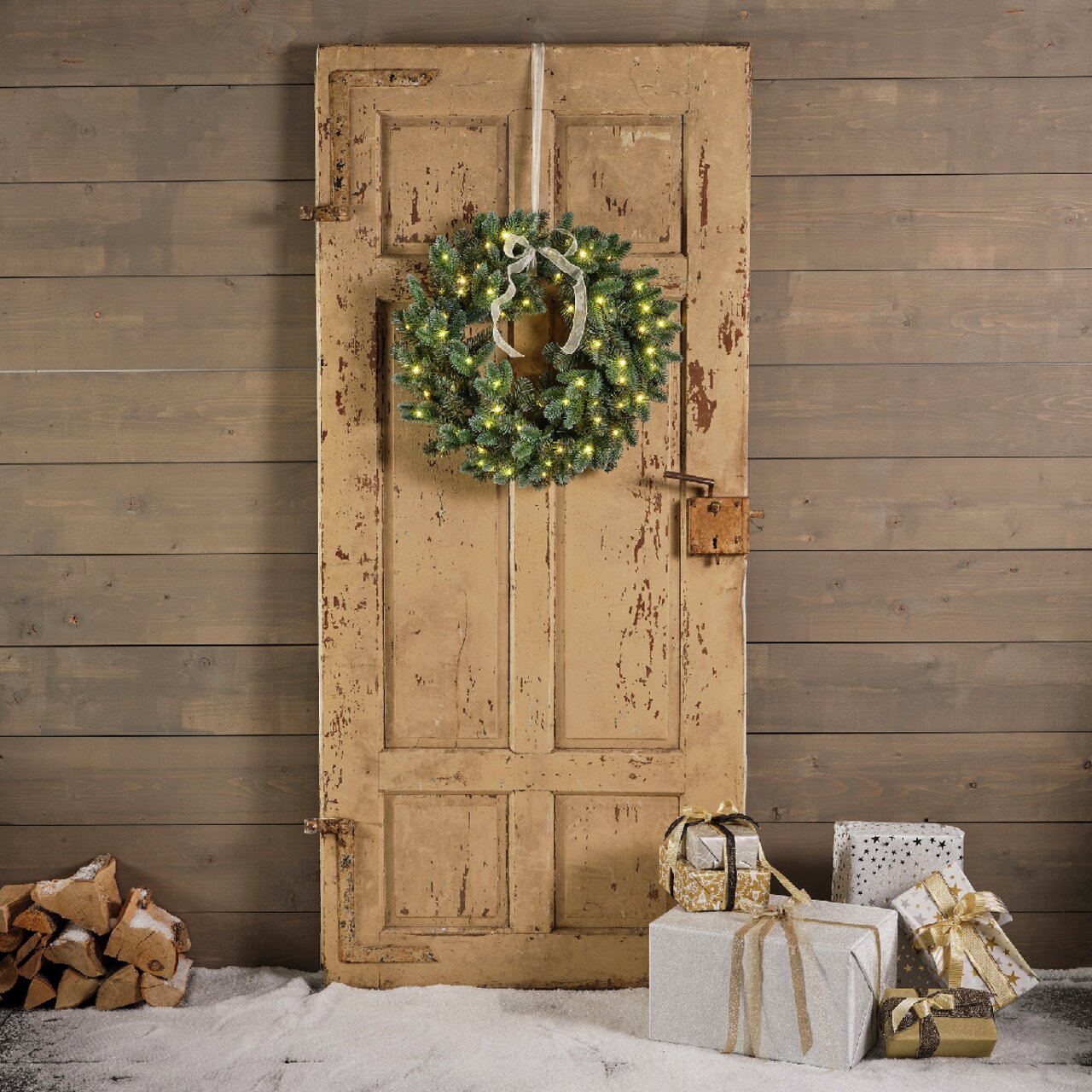 Weihnachtskranz mit Lichtern an einer alten Holztür, umgeben von Geschenken und Holzscheiten, aus frontaler Perspektive.