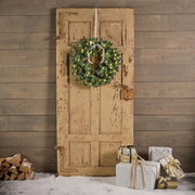 Weihnachtskranz mit Lichtern an einer alten Holztür, umgeben von Geschenken und Holzscheiten, aus frontaler Perspektive.