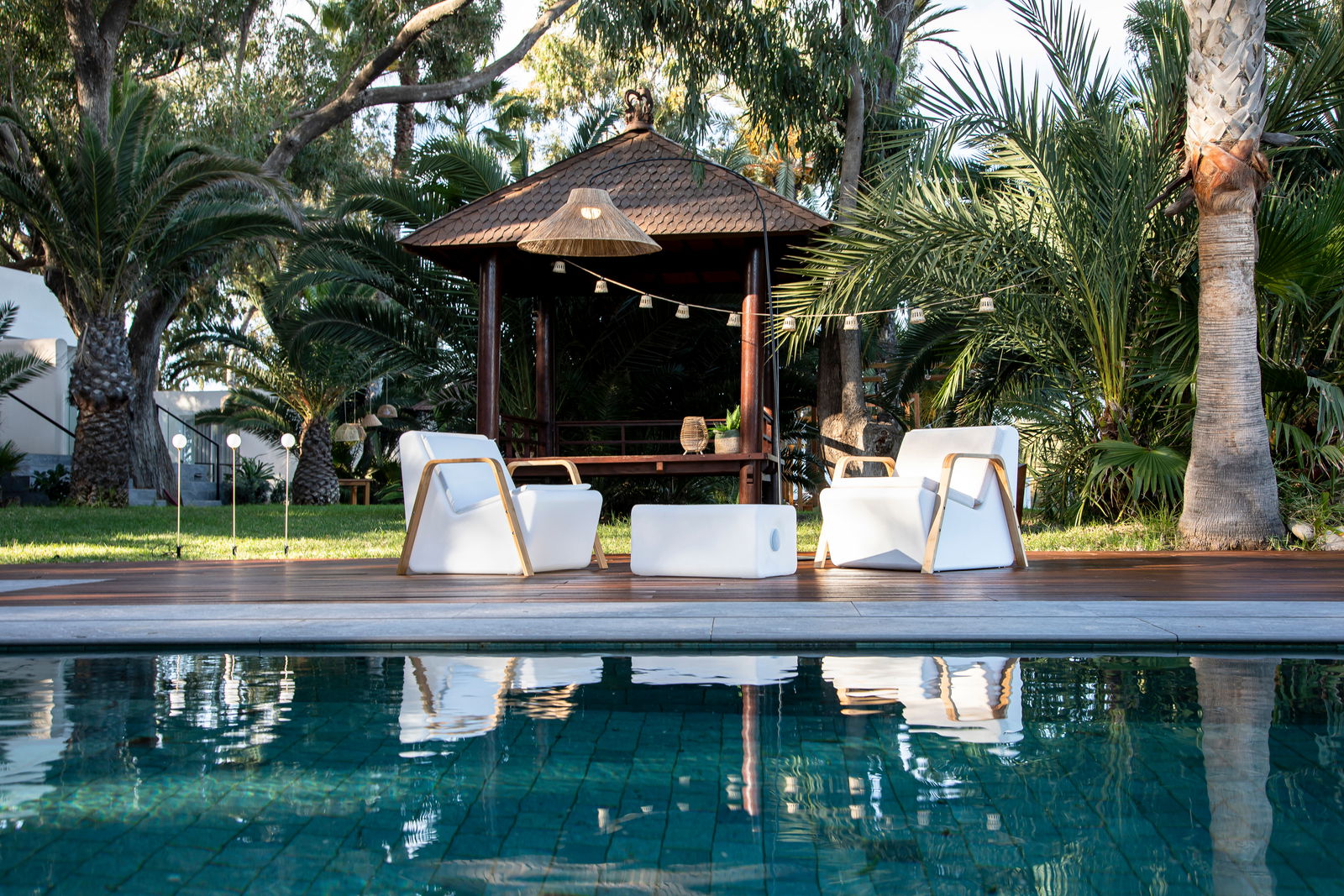Gartenansicht mit einem Pavillon im Hintergrund, umgeben von Palmen und grüner Vegetation. Im Vordergrund befinden sich moderne weiße Gartenmöbel auf einer Holzterrasse, die an einen Pool grenzt. Eine Girlande ist zwischen den Bäumen aufgehängt.