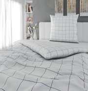 Weißes Bettwäsche-Set mit blauem Karomuster, Kissen und Decke auf einem Bett im Schlafzimmer, fotografiert in schräger Frontansicht von leicht oben.