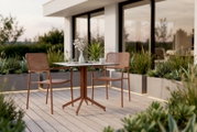 Zwei moderne Stapelsessel aus Metall in einem eleganten Terrakotta-Ton stehen auf einer stilvollen Terrasse mit Holzboden. Im Hintergrund sind Pflanzen und ein modernes Gebäude zu sehen. Die Perspektive zeigt die Sessel von der Seite.