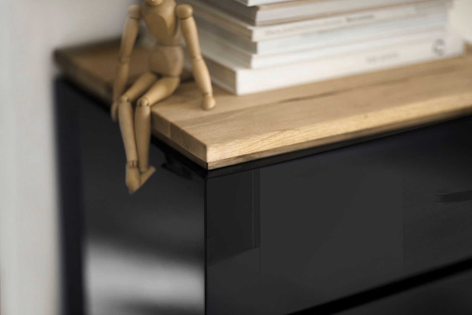 Nahaufnahme eines Möbelstücks mit schwarzer Hochglanzoberfläche und einer Holzplatte, darauf sitzt eine hölzerne Gliederpuppe neben einem Stapel Bücher. Perspektive von oben.