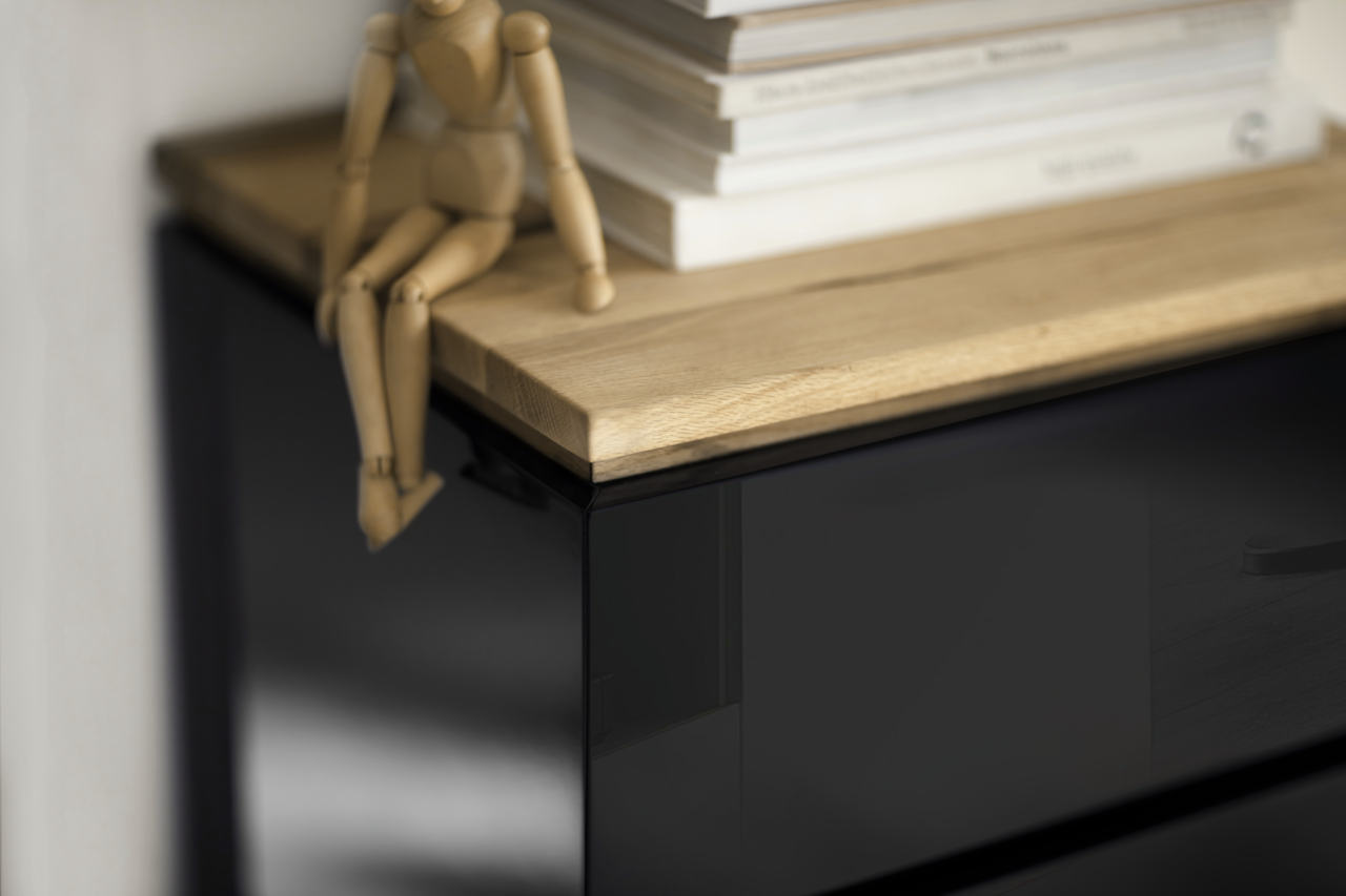 Nahaufnahme eines Möbelstücks mit schwarzer Hochglanzoberfläche und einer Holzplatte, darauf sitzt eine hölzerne Gliederpuppe neben einem Stapel Bücher. Perspektive von oben.