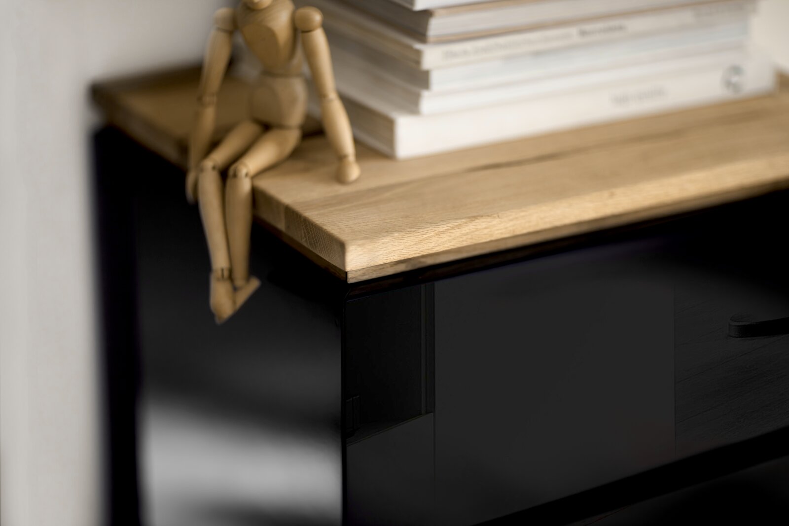 Nahaufnahme eines Möbelstücks mit schwarzer Hochglanzoberfläche und einer Holzplatte, darauf sitzt eine hölzerne Gliederpuppe neben einem Stapel Bücher. Perspektive von oben.
