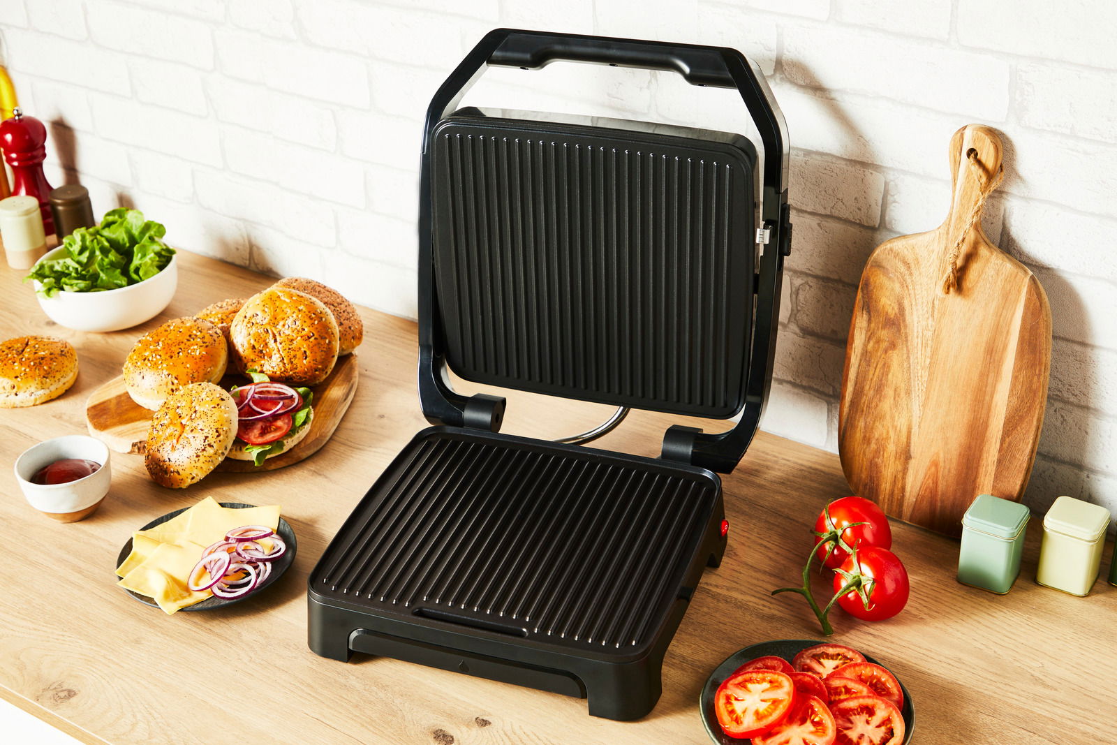 Kontaktgrill Classic GC271D in geöffneter Position auf einer Küchenarbeitsplatte, umgeben von Zutaten wie Brötchen, Salat und Tomaten, aus einer leicht seitlichen Perspektive.