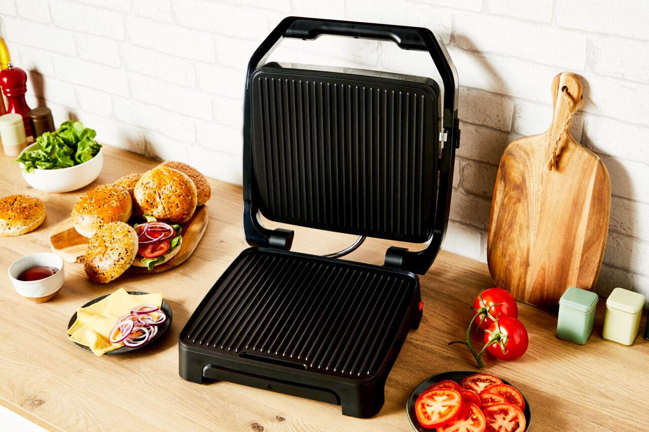 Kontaktgrill Classic GC271D in geöffneter Position auf einer Küchenarbeitsplatte, umgeben von Zutaten wie Brötchen, Salat und Tomaten, aus einer leicht seitlichen Perspektive.