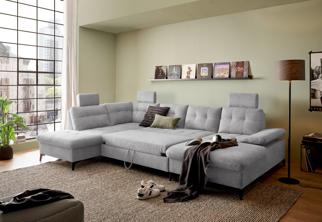 Graues Ecksofa in U-Form mit verstellbaren Kopfstützen, dekoriert mit Kissen, in einem modernen Wohnzimmer mit großen Fenstern und grüner Wandfarbe. Perspektive von vorne links.