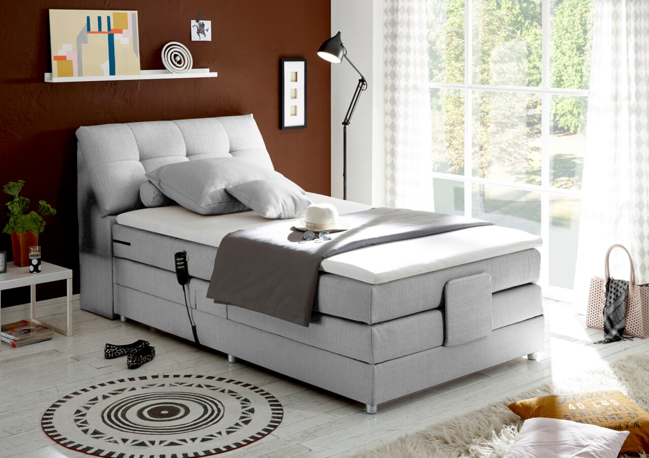 Graues Boxspringbett in modernem Schlafzimmer, seitliche Perspektive, mit Dekoration und Tageslicht.