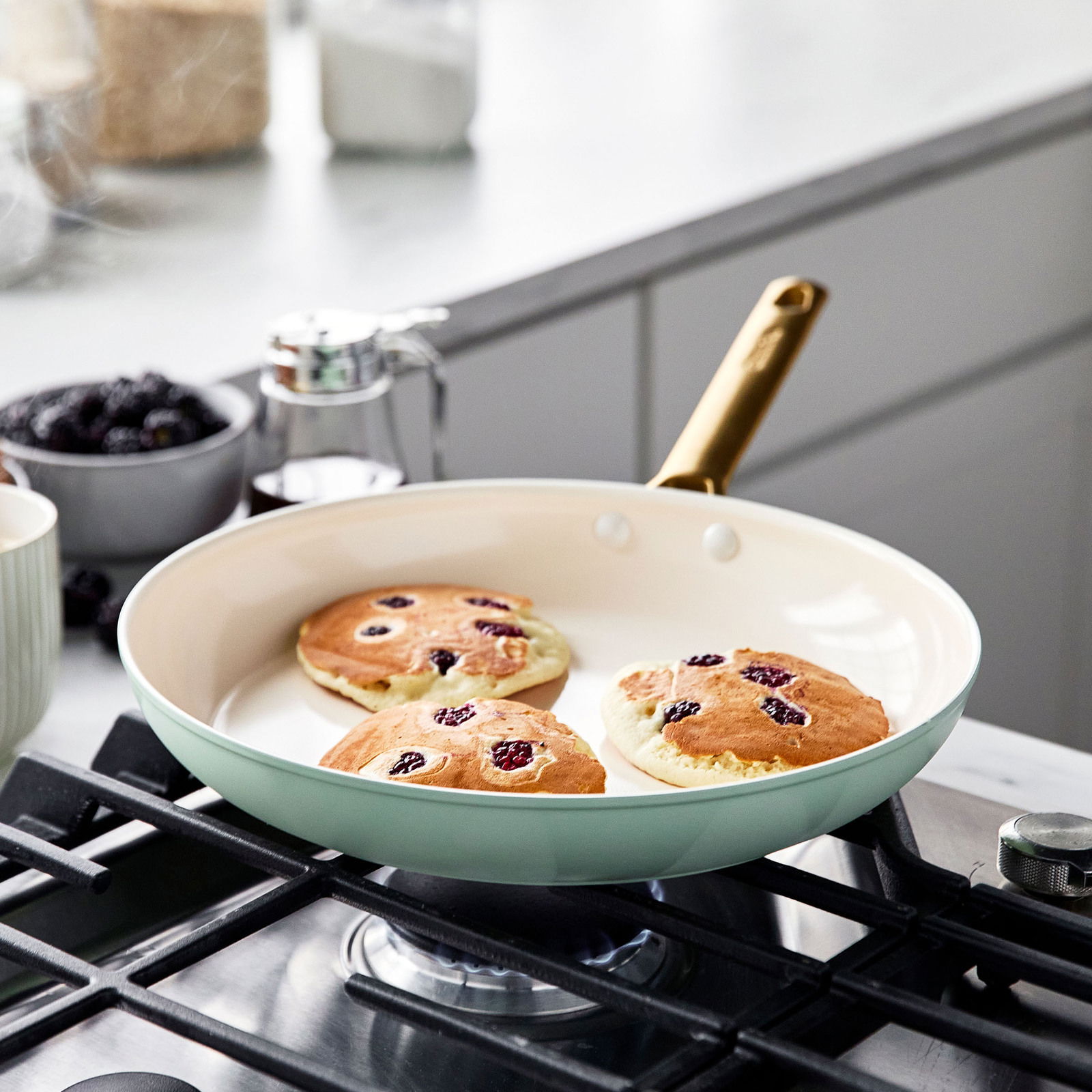 Pfanne mit goldfarbenem Griff auf einem Herd, in der drei Pfannkuchen mit Beeren gebacken werden, seitliche Perspektive.