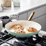Pfanne mit goldfarbenem Griff auf einem Herd, in der drei Pfannkuchen mit Beeren gebacken werden, seitliche Perspektive.