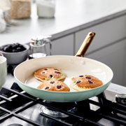 Een pan met een goudkleurig handvat op een fornuis, waarin drie pannenkoeken met bessen worden gebakken, gezien vanuit een zijaanzicht.