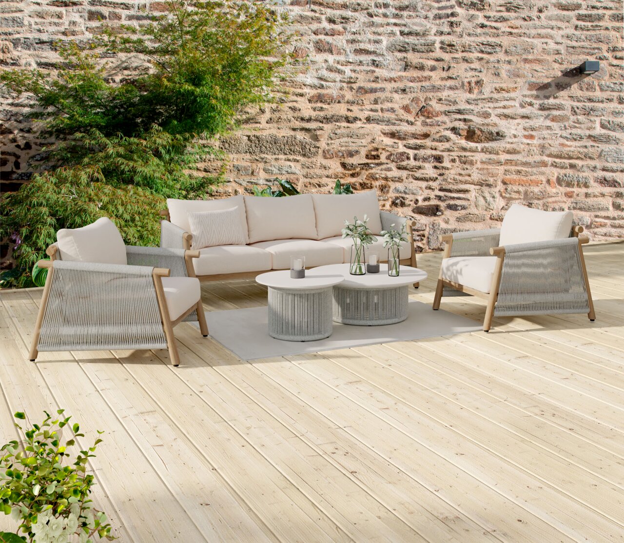 Outdoor-Lounge-Set auf Holzterrasse: beiges 3-Sitzer-Sofa mit zwei passenden Sesseln, grauen Seilbespannungen und zwei runden Couchtischen vor Steinmauer, aufgenommen in frontaler leicht seitlicher Perspektive.