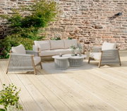 Outdoor-Lounge-Set auf Holzterrasse: beiges 3-Sitzer-Sofa mit zwei passenden Sesseln, grauen Seilbespannungen und zwei runden Couchtischen vor Steinmauer, aufgenommen in frontaler leicht seitlicher Perspektive.