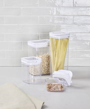 Vorratsdosen Set 8-tlg aus transparentem Kunststoff mit weißen Deckeln, gefüllt mit Spaghetti, Haferflocken und Nüssen, auf einer Küchenarbeitsplatte vor einer gefliesten Wand, seitliche Perspektive.