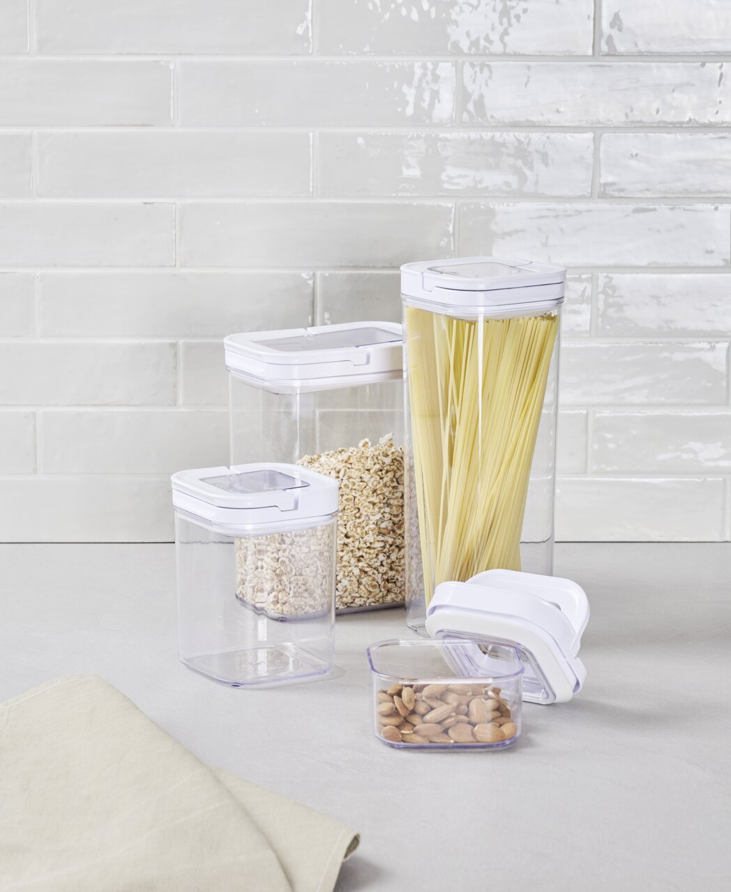 Homeware Set van 8 voorraadbussen Set van 8 voorraadbussen van doorzichtig kunststof met witte deksels, gevuld met spaghetti, havermout en noten, op een keukenblad voor een betegelde muur, zijaanzicht.