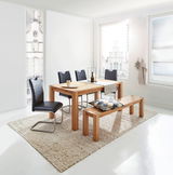 Esszimmer mit hellem Holztisch und passender Bank, umgeben von schwarzen Stühlen, in einem hellen Raum mit großen Fenstern, seitliche Perspektive.