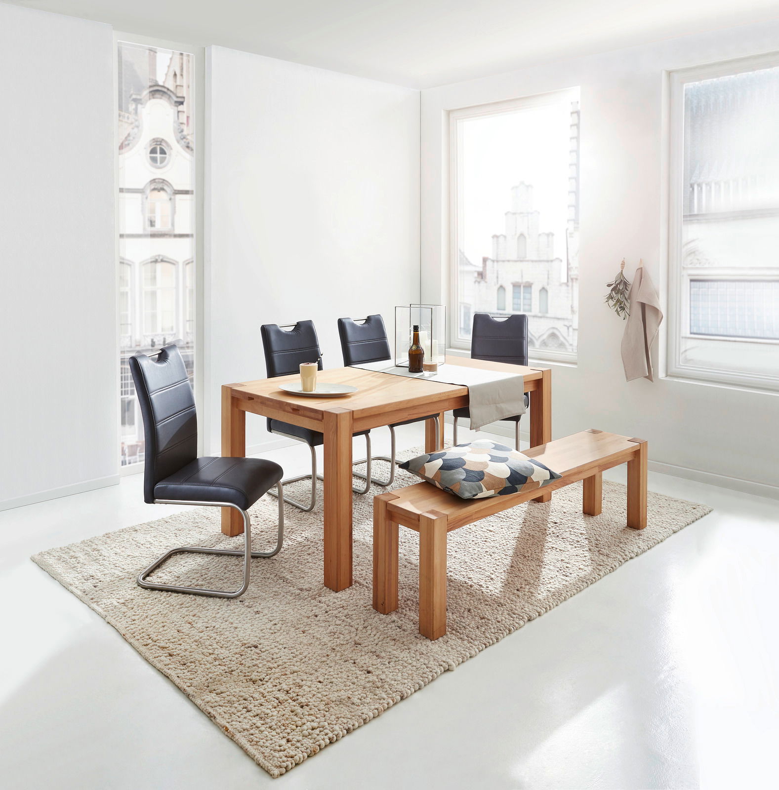 Esszimmer mit hellem Holztisch und passender Bank, umgeben von schwarzen Stühlen, in einem hellen Raum mit großen Fenstern, seitliche Perspektive.