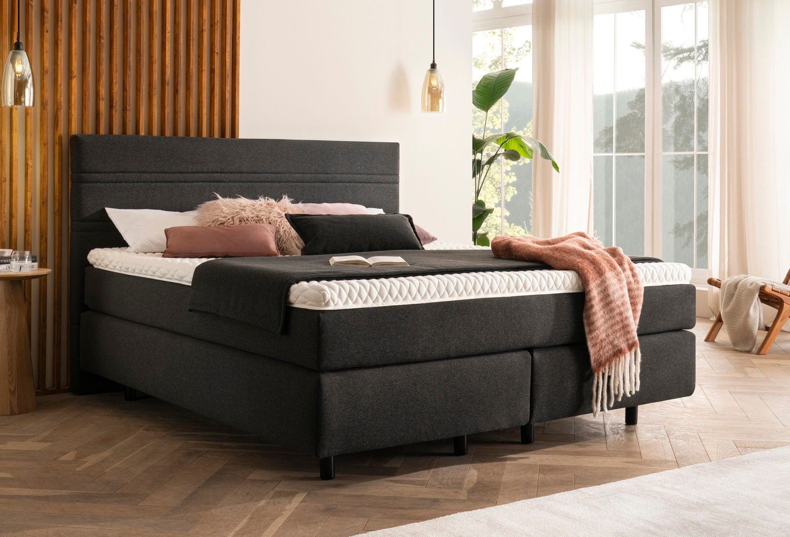 Boxspringbett in einem modernen Schlafzimmer, seitliche Perspektive, mit dunklem Stoffbezug, dekoriert mit Kissen und Decke, vor großen Fenstern.
