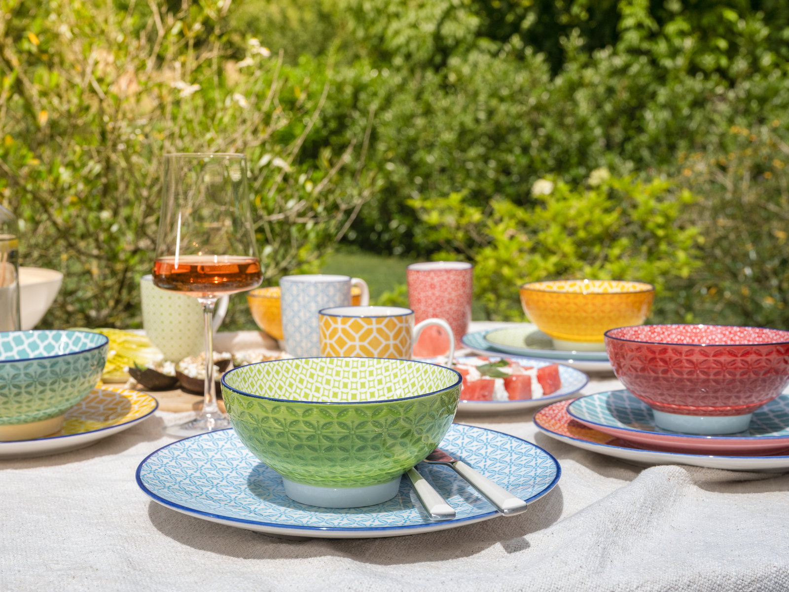 Bunte Geschirrkollektion im Freien, bestehend aus Schalen und Tellern in verschiedenen Farben und Mustern, mit einem Glas Roséwein im Vordergrund.