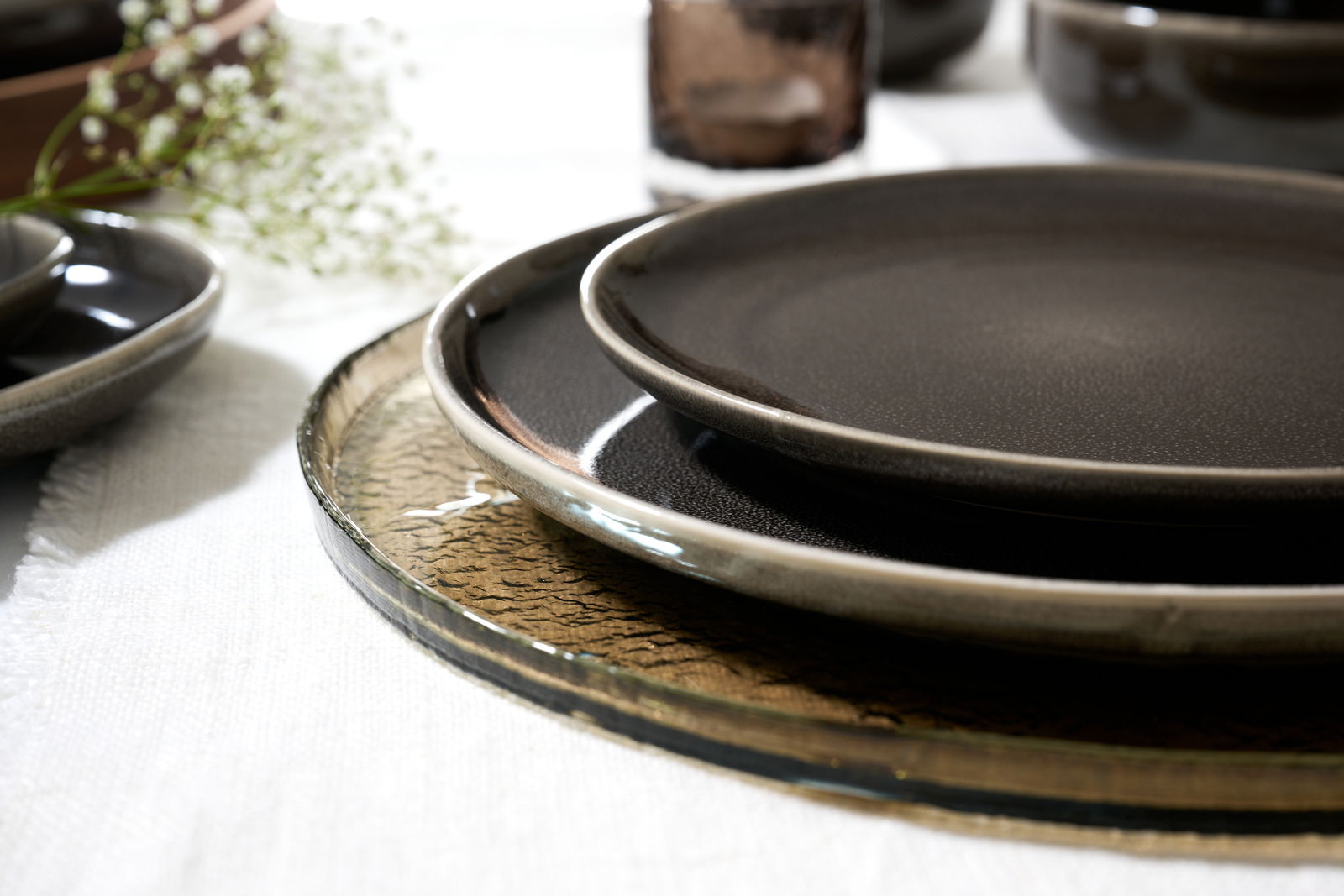 Ritzenhoff & Breker Beilagenteller EDA UMBRA Nahaufnahme von gestapelten, dunklen Tellern auf einem weißen Tischtuch, mit einem unscharfen Hintergrund aus weiteren Geschirrteilen und Blumen.