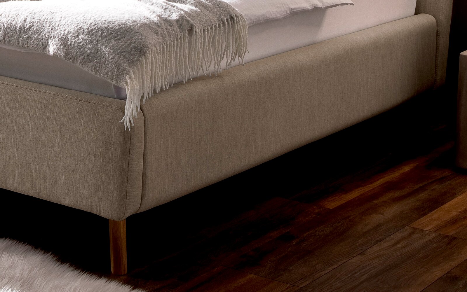 Nahaufnahme eines Polsterbetts mit beigem Stoffbezug und Holzfüßen, seitliche Perspektive