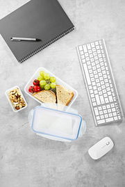 Frischhaltedosen-Set aus der Vogelperspektive mit belegtem Sandwich, Trauben, Tomaten und Nüssen, neben einem Notizbuch, Stift, Tastatur und Maus auf einem Schreibtisch.