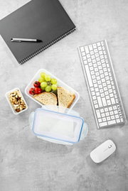 Frischhaltedosen-Set aus der Vogelperspektive mit belegtem Sandwich, Trauben, Tomaten und Nüssen, neben einem Notizbuch, Stift, Tastatur und Maus auf einem Schreibtisch.