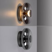 Moderne Wandleuchte mit rauchgrauem, kuppelförmigem Glasschirm und rundem Metall-Wandhalter, warm leuchtend, seitliche Ansicht (Profil) an heller Wand.
