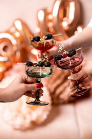 Drei Sekt- und Dessertschalen in verschiedenen Farben, gefüllt mit Beeren und dekoriert mit Blumen, werden von Händen in einer festlichen Atmosphäre gehalten. Perspektive: Nahaufnahme von oben.