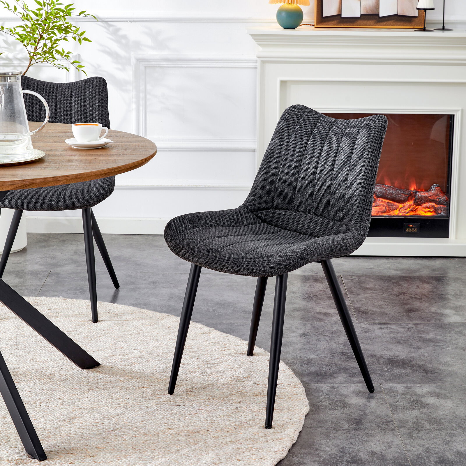 Dunkelgrauer, gepolsterter Esszimmerstuhl mit Stoffbezug und schwarzen Metallbeinen neben rundem Holztisch im Wohnbereich, perspektivisch in schräger Vorderansicht von leicht oben.
