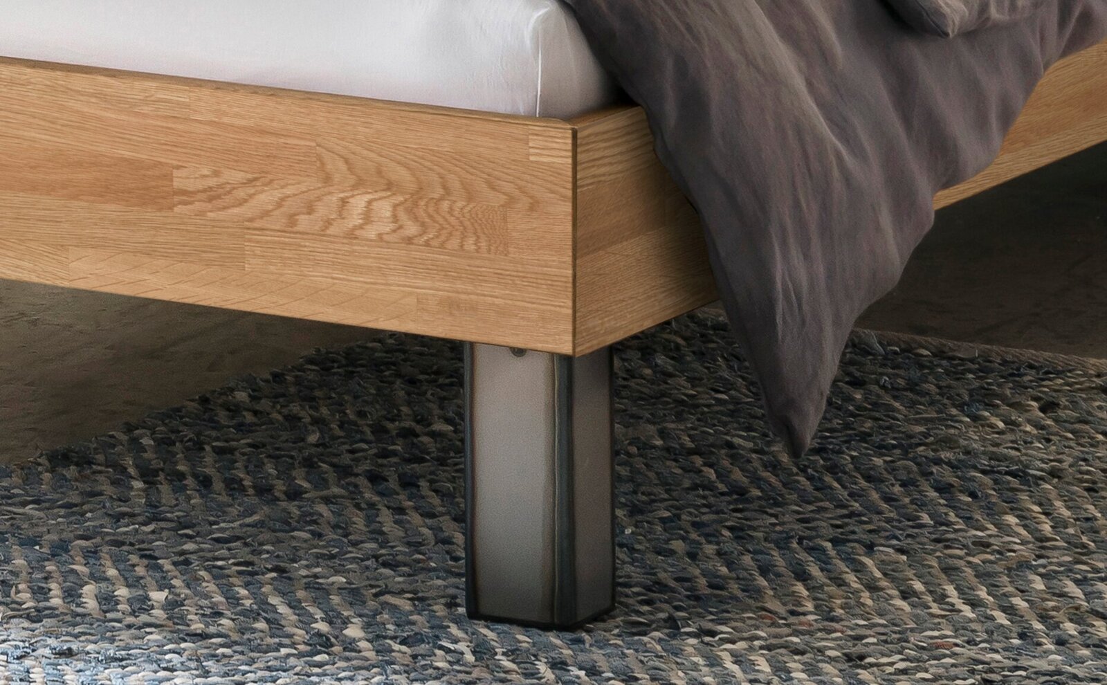 Detailansicht eines Bettrahmens aus hellem Holz mit Metallfuß und grauer Bettwäsche auf Teppich, seitliche Perspektive auf die Ecke des Bettes