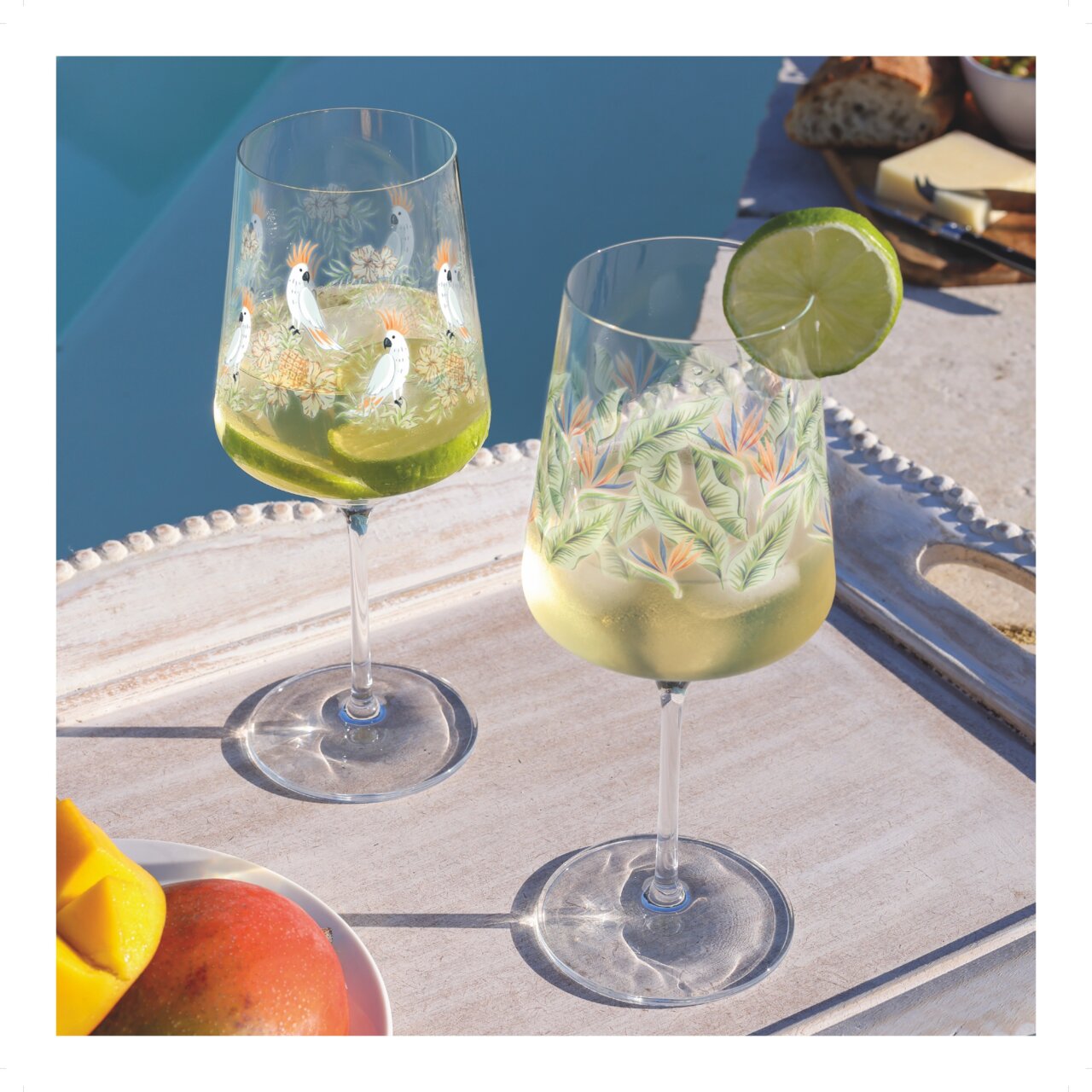 Zwei dekorative Weingläser mit tropischen Motiven, gefüllt mit einem erfrischenden Getränk und Limettenscheiben, auf einem Tablett am Pool bei Tageslicht.