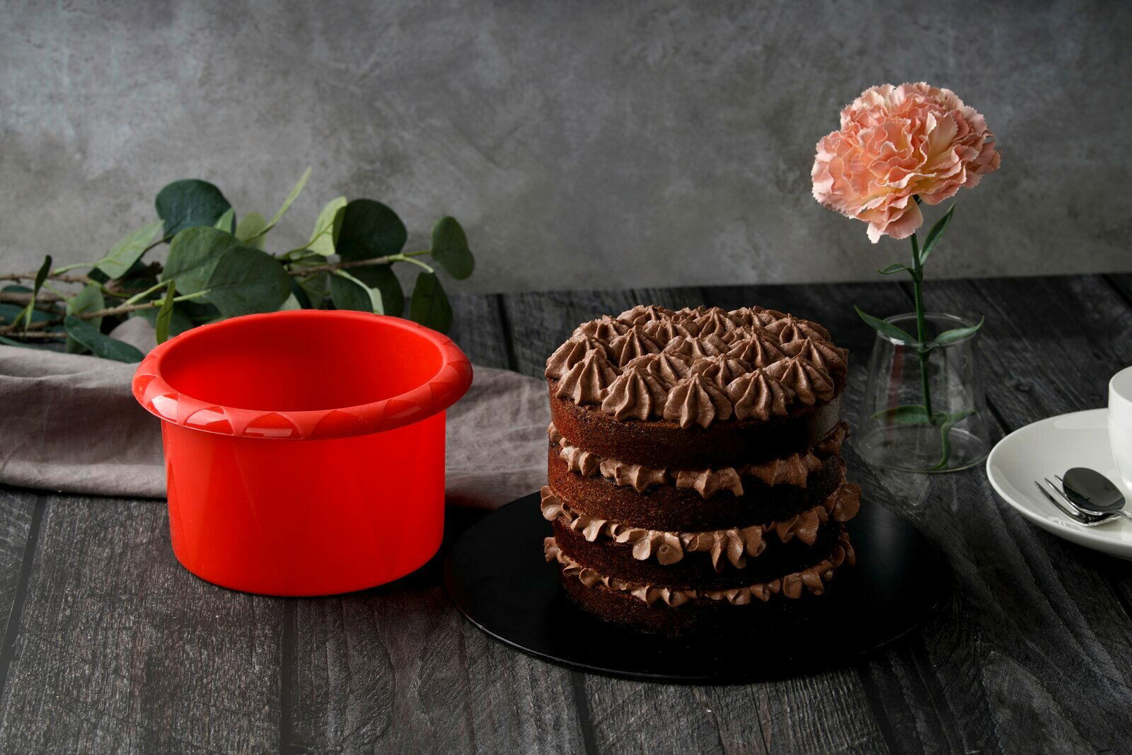 Rote Silikon-Rundform mit einem Durchmesser von 16 cm, neben einer mehrschichtigen Schokoladentorte, aus seitlicher Perspektive fotografiert.