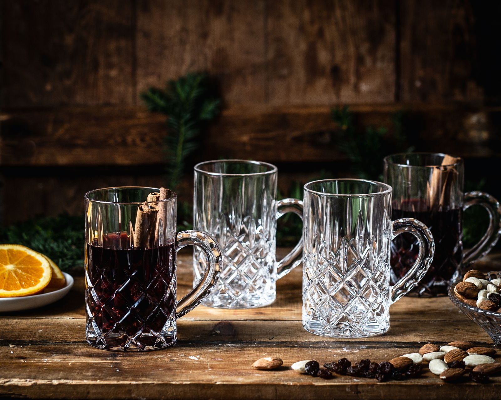Vier elegante Glühwein-Gläser Noblesse mit Henkel, zwei gefüllt mit Glühwein und Zimtstangen, auf einem rustikalen Holztisch mit Nüssen und Orangenscheiben, seitliche Perspektive.
