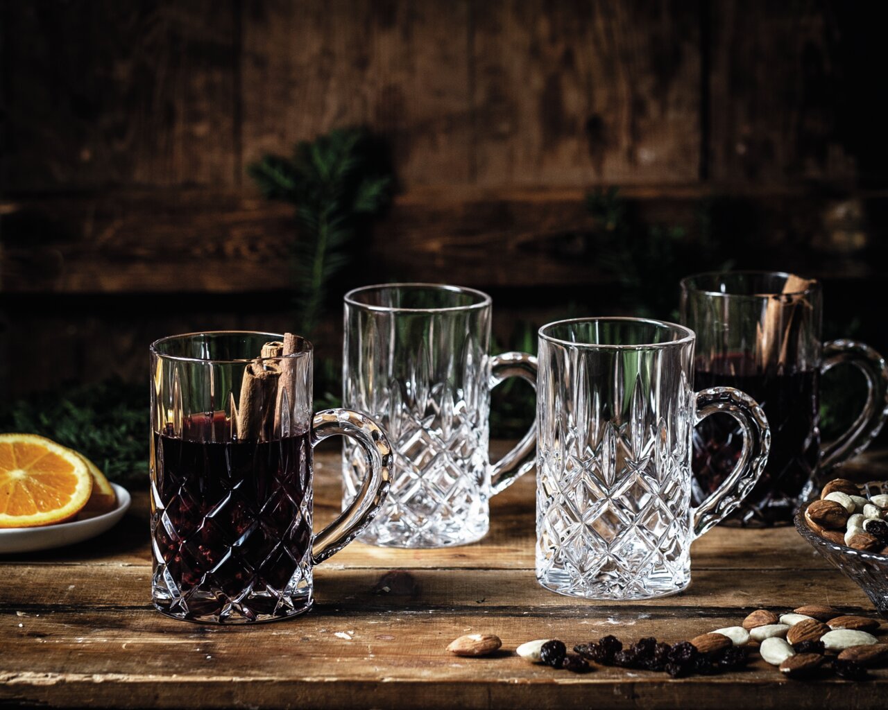 Vier elegante Noblesse-glühweinglazen met oor, waarvan er twee gevuld zijn met glühwein en kaneelstokjes, op een rustieke houten tafel met noten en schijfjes sinaasappel, zijaanzicht.