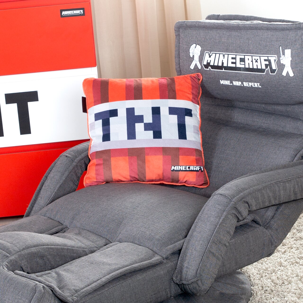 Zurbrüggen Dekokissen TNT 38x38cm MINECRAFT Grauer Minecraft-Gaming-Sessel mit Armlehnen und gesticktem Minecraft-Logo auf der Rückenlehne, mit TNT-Dekokissen auf der Sitzfläche, schräg von vorne fotografiert.