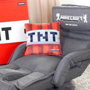 Grauer Minecraft-Gaming-Sessel mit Armlehnen und gesticktem Minecraft-Logo auf der Rückenlehne, mit TNT-Dekokissen auf der Sitzfläche, schräg von vorne fotografiert.