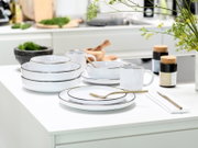 Weißes Geschirr-Set mit dunklem Rand (Teller, Schalen und Becher) auf weißer Kücheninsel mit goldenem Besteck, fotografiert aus leicht erhöhter Frontperspektive