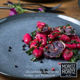 Nahaufnahme eines blauen Tellers mit einem kunstvoll angerichteten Gericht aus roten Gnocchi, roten Zwiebeln und Kräutern, dekoriert mit schwarzen und roten Pfefferkörnern. Der Teller liegt auf einem Holztisch, im Hintergrund sind unscharf Besteck und Blu