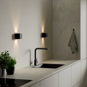 Moderne kubische LED-Wandleuchte in Schwarz mit Up-&-Down-Light an heller Küchenwand über Spüle, perspektivisch in leichter Seitenansicht