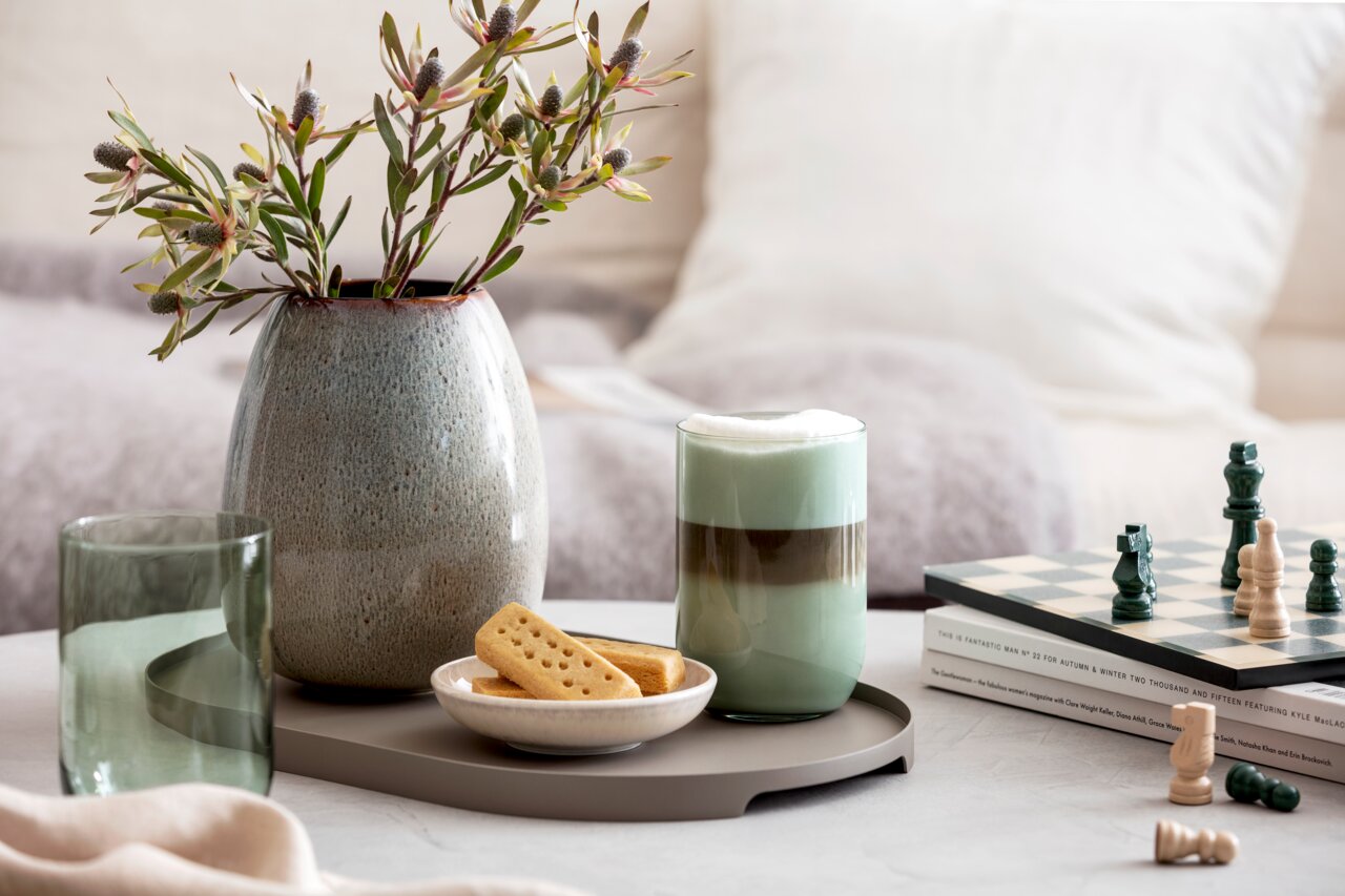 Like by Villeroy & Boch Longdrinkbecher Set 2-tlg. LIKE SAGE Ein stilvolles Longdrinkbecher-Set auf einem Tisch, umgeben von einer Vase mit Blumen, einem Schachbrett und einem Teller mit Keksen. Perspektive von vorne.