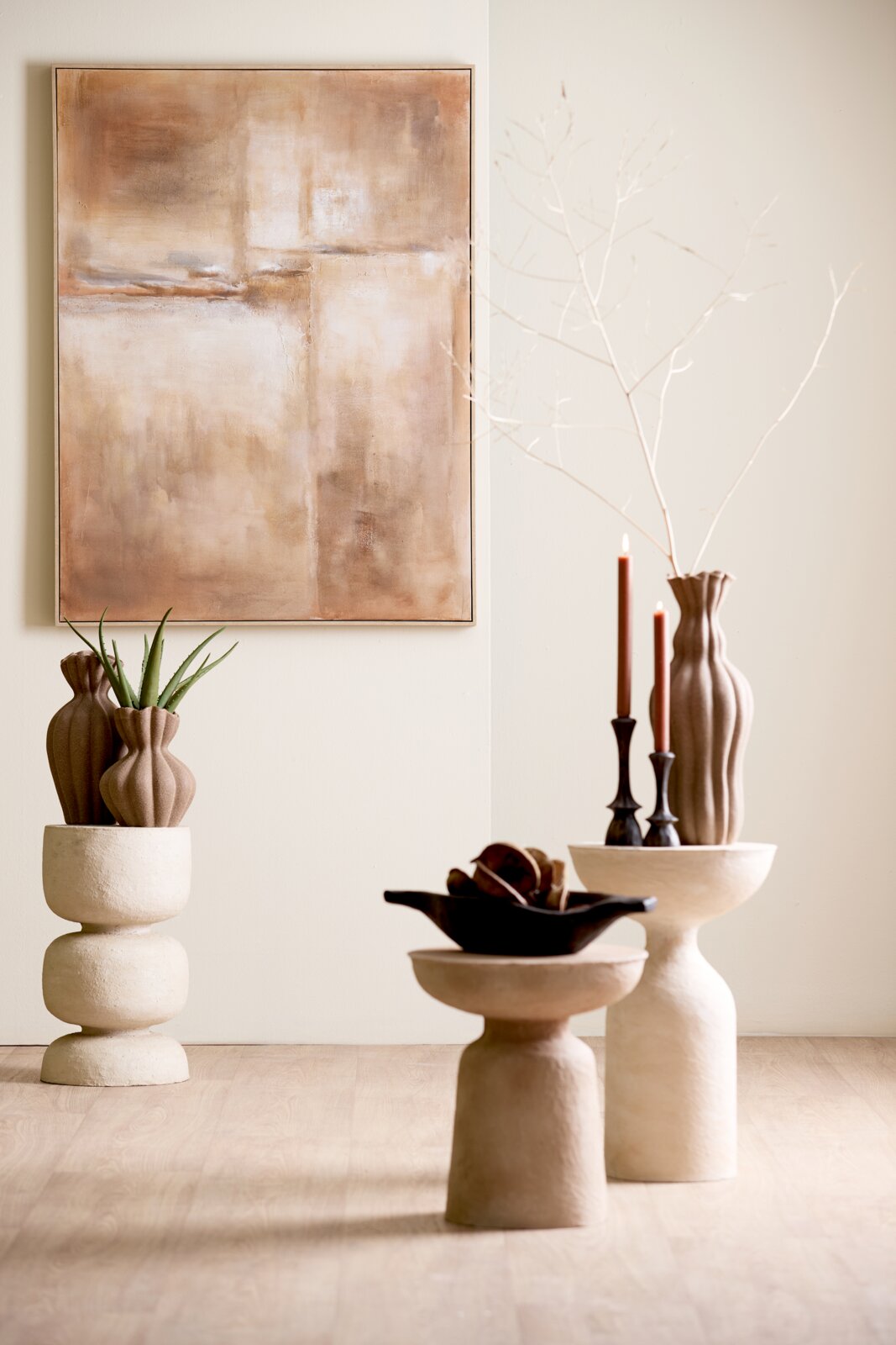 Vooraanzicht van een warm, minimalistisch interieur met abstract schilderij, sculpturale bijzettafels, organische vazen, brandende kaarsen en een aloëplant in natuurlijke beige- en bruintinten.
