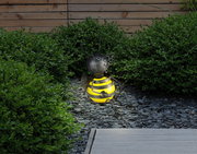 Niedliche Solarleuchte in Form einer Biene, die auf einem Kiesweg im Garten sitzt, umgeben von grünen Büschen, seitliche Perspektive.