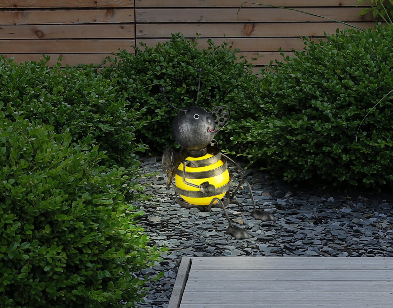 Niedliche Solarleuchte in Form einer Biene, die auf einem Kiesweg im Garten sitzt, umgeben von grünen Büschen, seitliche Perspektive.