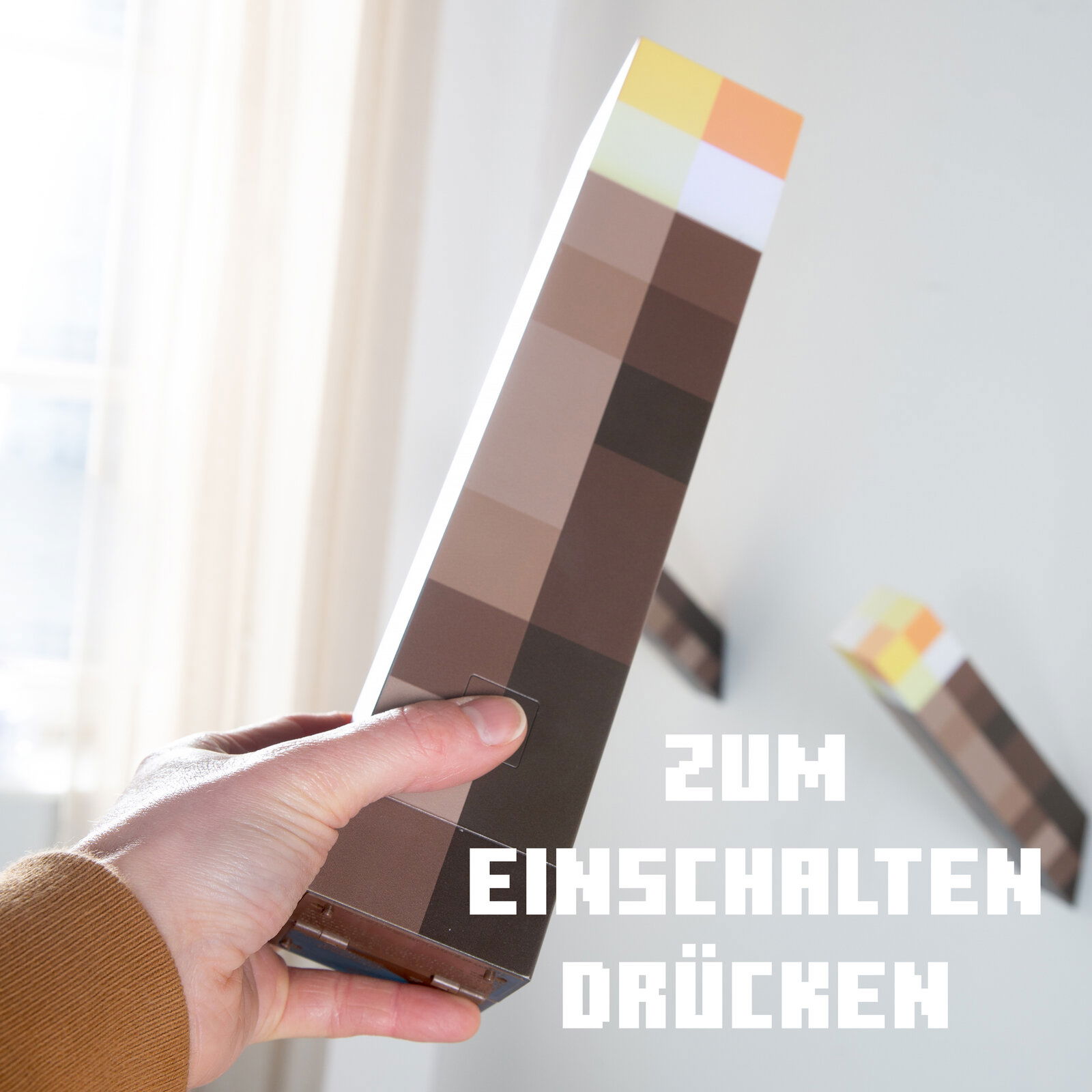 Hand hält eine pixelige Minecraft-Fackel-Lampe mit sichtbarem Einschaltknopf, schräg von vorn im Innenraum fotografiert; Text im Bild: „ZUM EINSCHALTEN DRÜCKEN“.