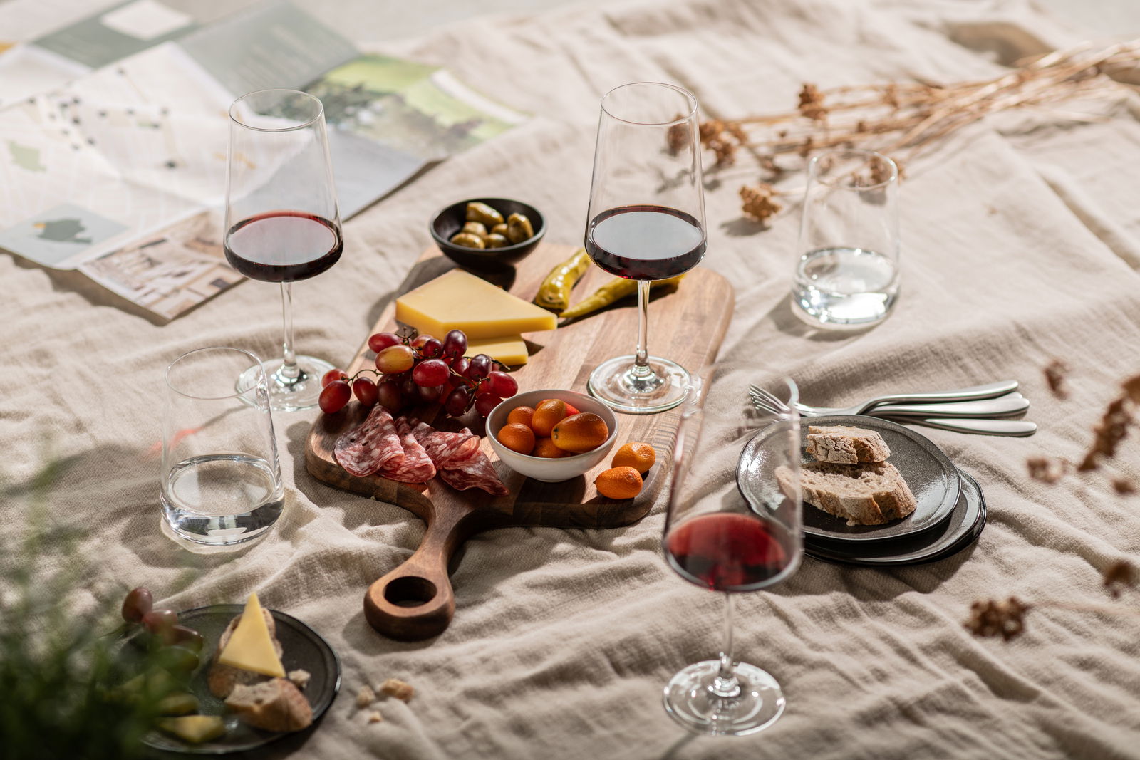 SCHOTT ZWIESEL Becher 4er-Set MIOVINO Ein stilvolles Picknick-Arrangement mit einem Becher-Set, bestehend aus Weingläsern und Wassergläsern, auf einer Decke. Im Vordergrund sind Käse, Trauben, Salami und Brot auf einem Holzbrett zu sehen. Perspektive von oben.