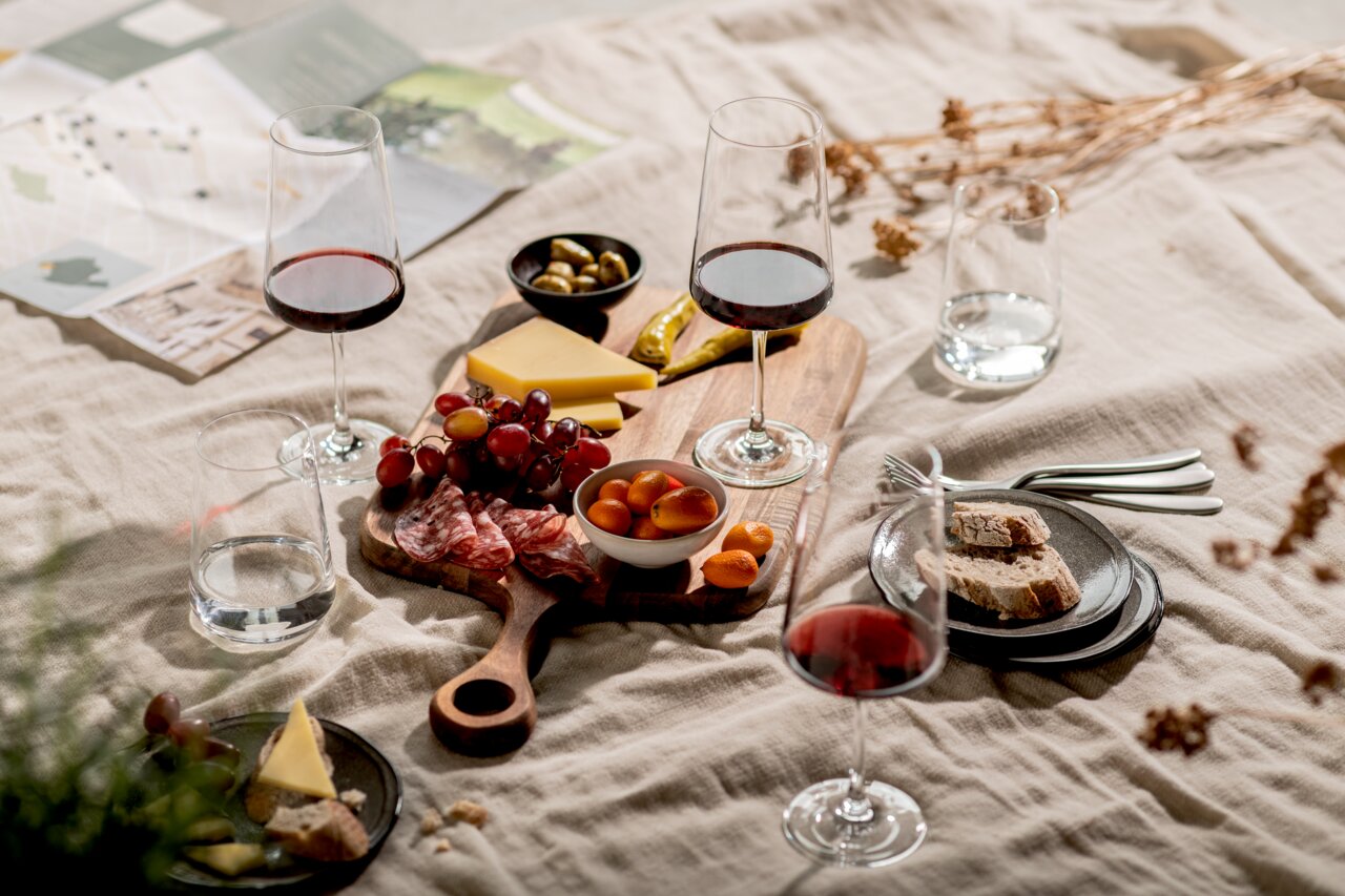 Ein stilvolles Picknick-Arrangement mit einem Becher-Set, bestehend aus Weingläsern und Wassergläsern, auf einer Decke. Im Vordergrund sind Käse, Trauben, Salami und Brot auf einem Holzbrett zu sehen. Perspektive von oben.